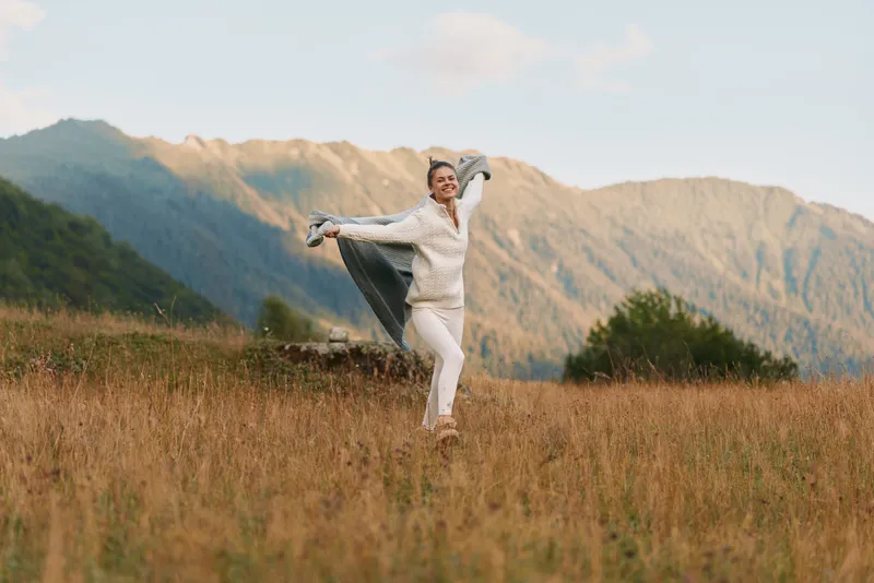 Joyful woman in white dances across a grassy field with mountains in the background, conveying a bright outdoor lifestyle moment, warm natural light, and freedom.