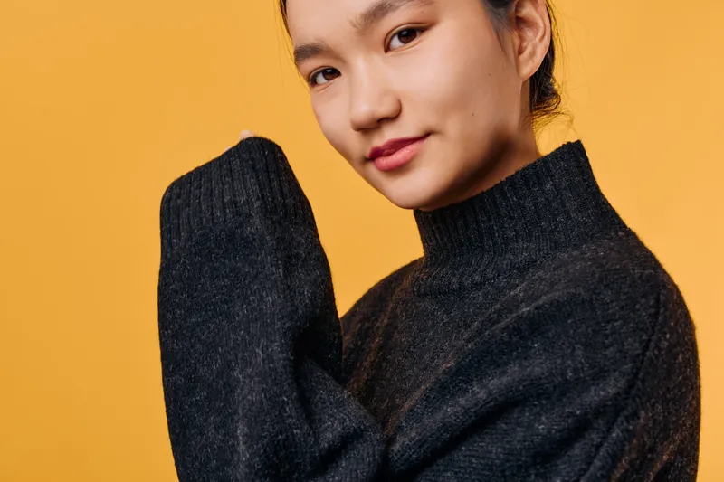 Young woman with black sweater posing on yellow background smiling gently with relaxed face and casual posture, studio portrait of confident female model looking at camera.