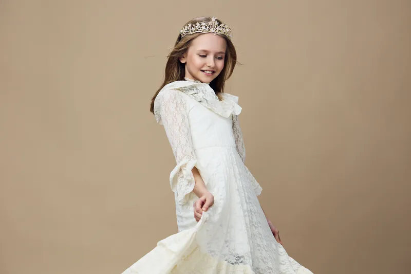 Young girl in a white princess dress smiling with a crown, set against a soft beige background, exuding joy and elegance, perfect for children s fashion themes