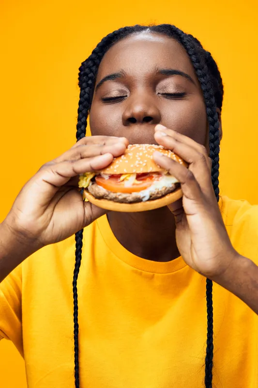 Young Black woman enjoying a delicious hamburger against a vibrant yellow background, showcasing a joyful expression and casual attire, emphasizing delicious fast food