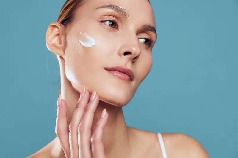Close up of a young woman applying white cream to her cheek, a calm skincare moment against a blue background, showcasing self care and focus.