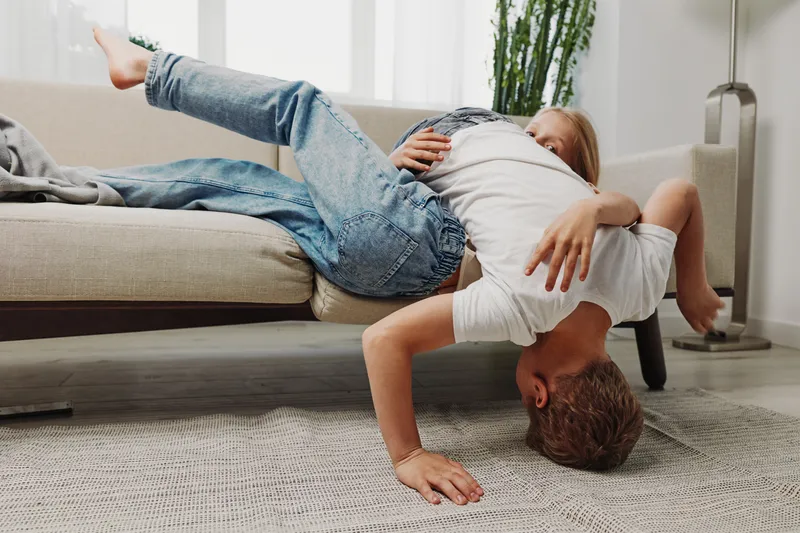Two children playfully wrestle on the floor near a sofa in a bright living room, showing fun and joyful family interaction with casual clothing.
