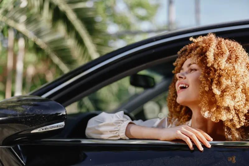 smiling woman with curly hair enjoying summer drive in black car, tropical background, carefree and happy mood, bright natural light, outdoor lifestyle moment