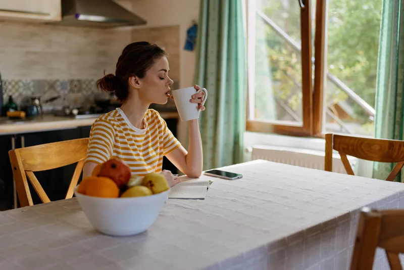A woman sits at a bright kitchen table sipping from a mug, fruit bowl in the foreground. A simple home lifestyle scene focused on morning routine, calm, and comfort.