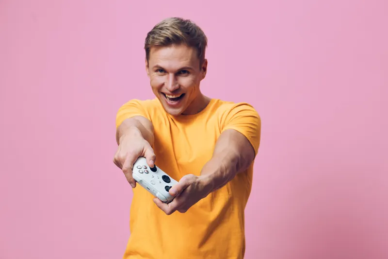 Smiling man wearing bright yellow t-shirt playfully holding game controller towards camera on pink background studio portrait. People entertainment concept