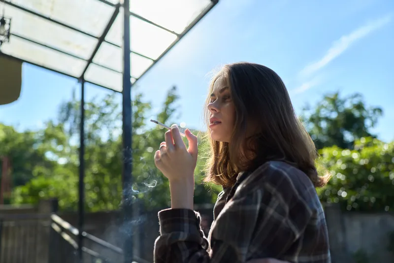 A woman in plaid casual attire stands outdoors, holding a small object near her mouth, sunlight highlighting a contemplative, natural expression in a relaxed lifestyle moment
