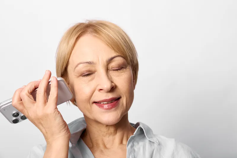 Elderly woman portrait talking on smartphone during call, calm relaxed expression, close view, light gray studio background, mobile communication concept, wireless connection, copy space