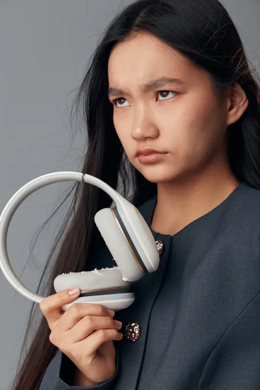 Young woman holding broken headphones with visible damage on ear pads showing frustration and disappointment. Portrait with serious expression on neutral background wearing dark outfit indoors.