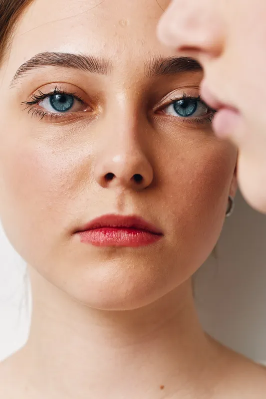 Closeup portrait of young woman with blue eyes and natural skin looking at camera, another person profile blurred in foreground, studio lighting and neutral background.