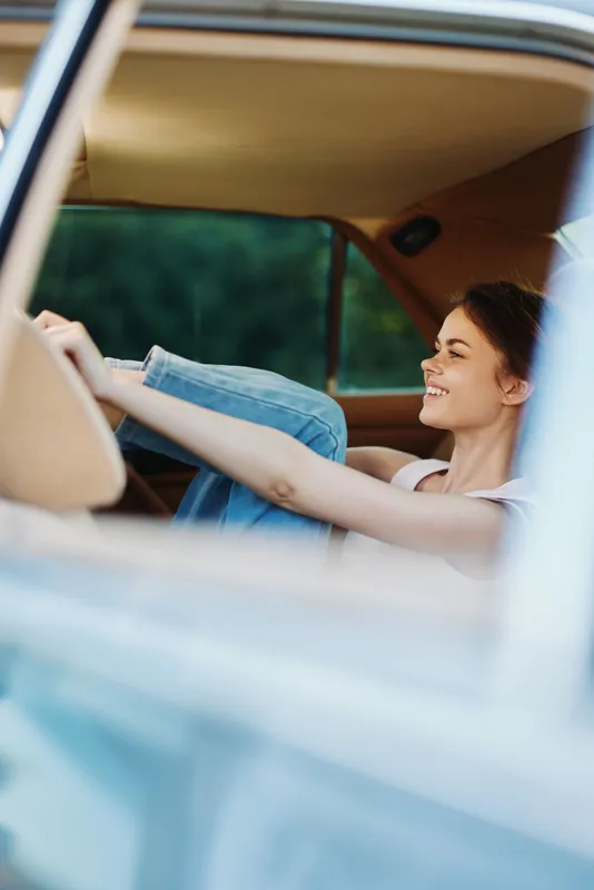 Young woman enjoying her joyful moment while sitting in a vintage car, showcasing a carefree spirit and casual denim fashion against a natural green background.
