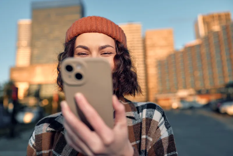Woman taking a selfie with smartphone in urban street at golden hour, candid smile wearing beanie, authenticity and mindful living captured in emotional storytelling and candid lifestyle.