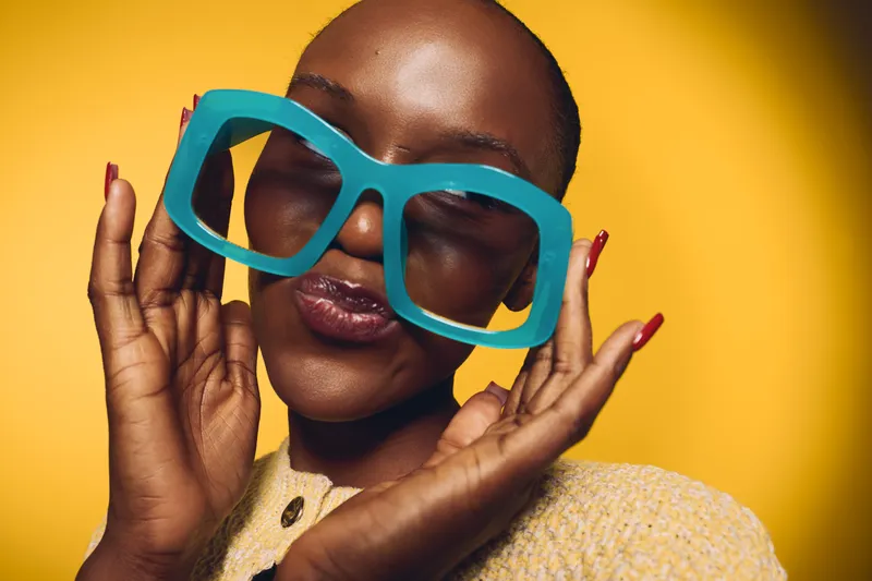 Happy black woman with dark skin wearing oversized blue glasses smiles with closed eyes on yellow background. Concept of joy, fashion, and playful mood in studio portrait.