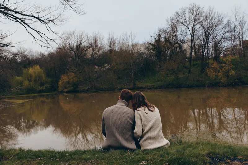 A couple sits close by a calm lake in autumn, wearing cozy sweaters, sharing a quiet moment, soft breeze, muted colors, reflective water, peaceful countryside setting