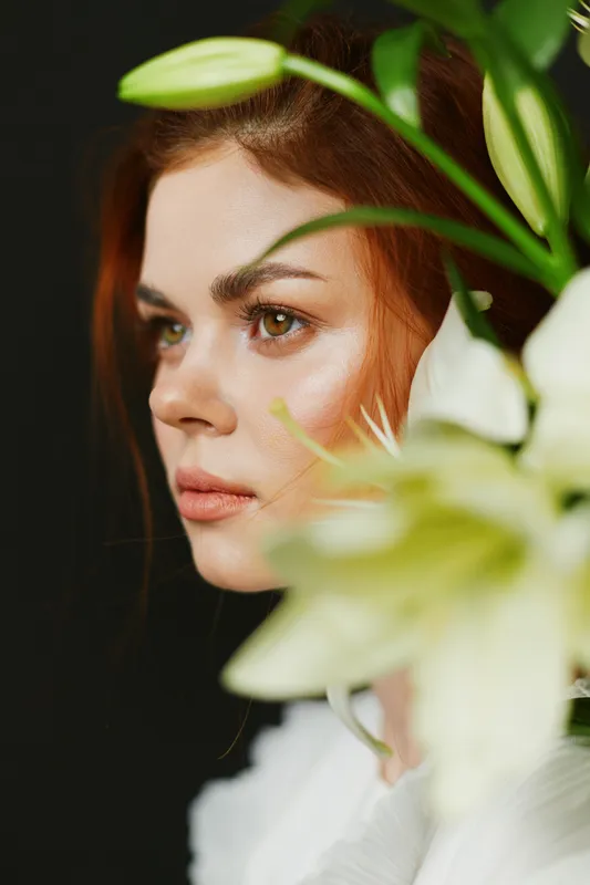 Elegant portrait of a young woman with red hair, featuring white lilies in a dark background, expressing serenity and beauty in a seasonal spring theme