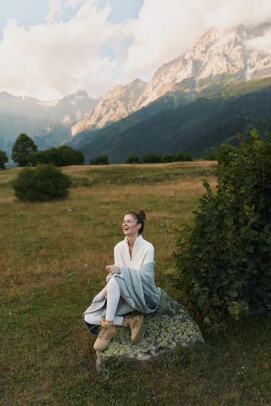 Young woman lifestyle enjoying nature sits on rock wrapped in blanket smiles relaxed with mountain landscape background under cloudy sky in countryside outdoors.