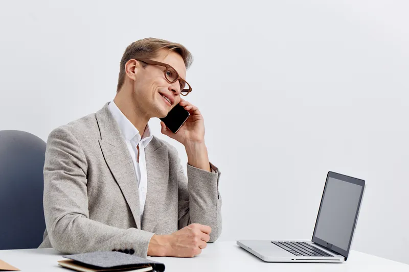 Smiling professional man talking on mobile phone while working at office desk with laptop and notepad, business casual attire, light background, positive mood concept, modern workplace.