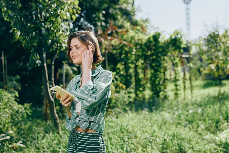 Young woman stands outdoors in a park, wearing a striped shirt and dress, using a smartphone amid sunlit greenery.