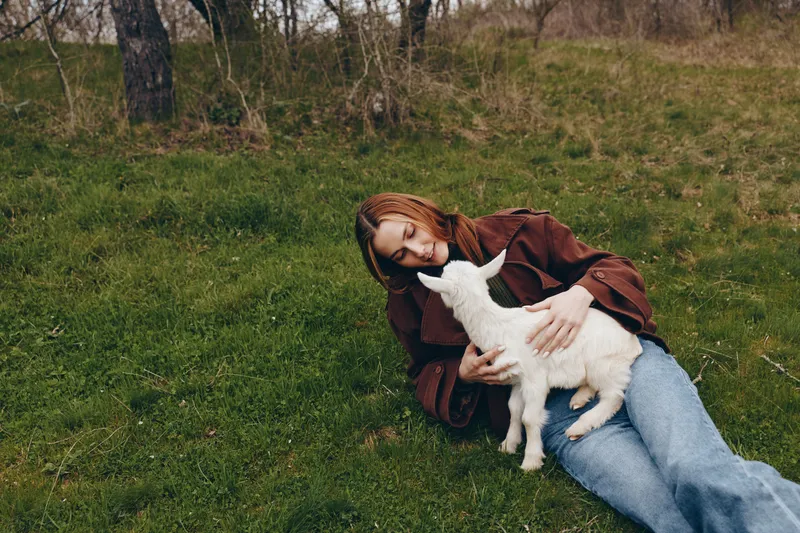 Lifestyle moment outdoors with a woman resting on grass, sharing gentle affection with a kid goat, natural colors, serene meadow scene, warm jacket, soft breeze, candid pose, rustic charm