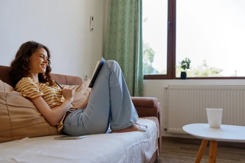 A young woman lounges on a sofa at home, smiling while browsing on a tablet in a bright cozy living room with soft sunlight and a cup nearby.
