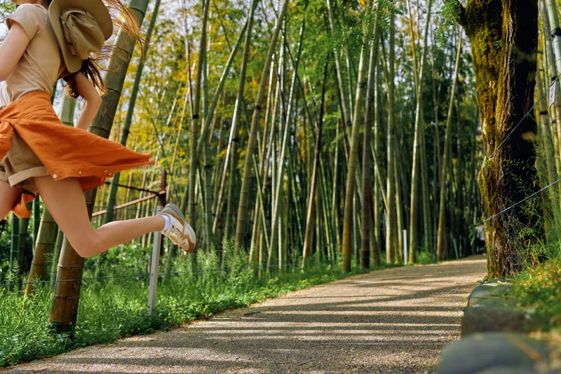 Girl jump on pathway in bamboo park with sun hat and orange skirt, summer motion captured midair, carefree child enjoying gravel trail under tall green bamboo trees and sunlight.