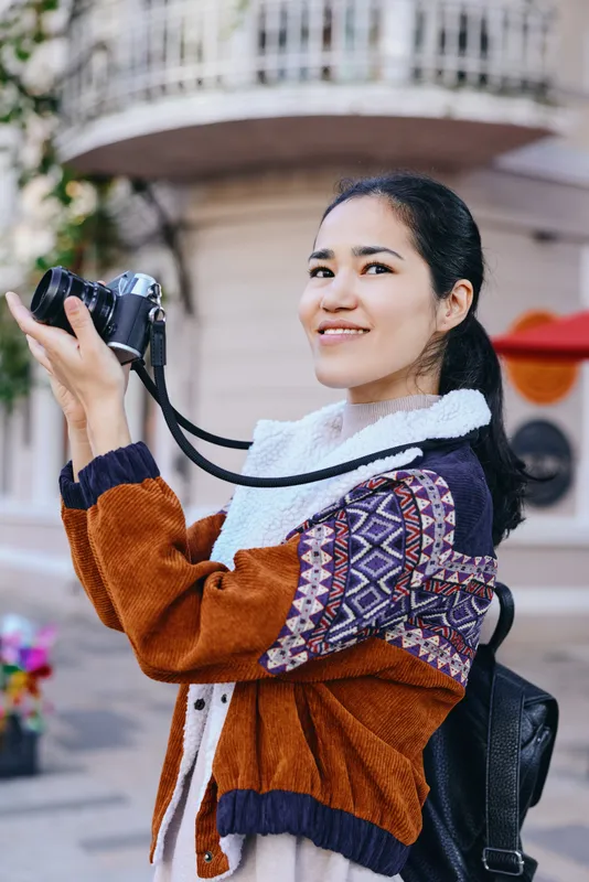Candid shot of a woman photographing with a camera on a sunny street, wearing a patterned cardigan and backpack, showcasing outdoor photography, travel moments, joyful exploration, and creative