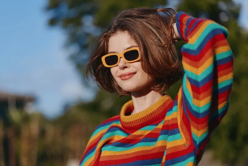 Woman in a rainbow sweater and yellow sunglasses smiling confidently outdoors, short brown hair tucked back with hand in hair, casual colorful fashion portrait for lifestyle.