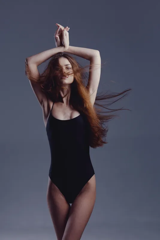 Smiling woman with long hair in black bodysuit dancing against gray background with flowing hair and relaxed expression.