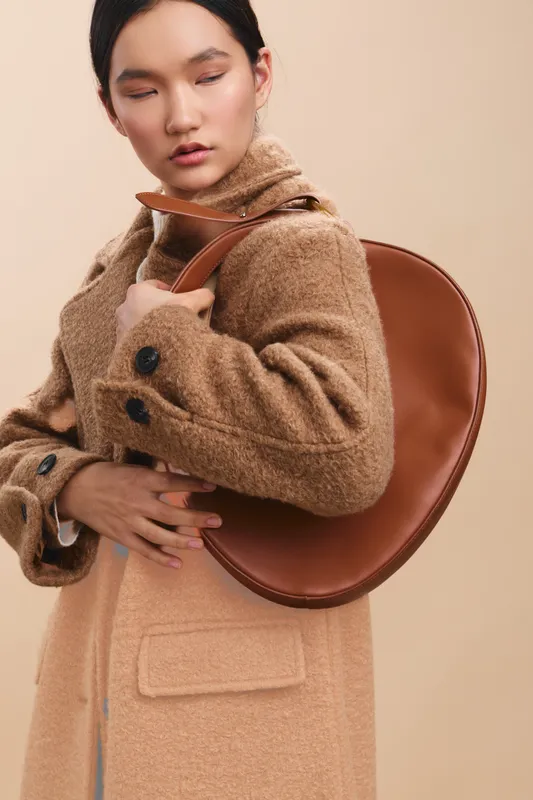 asian woman wearing a beige wool coat holds a brown handbag close, posing in a studio with soft lighting, exuding elegance, warmth, and contemporary fashion sophistication.