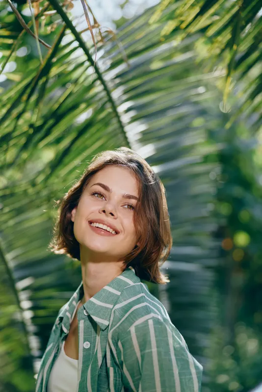 Bright portrait of a joyful woman in a sunny tropical setting, wearing a striped green shirt, framed by lush palm leaves.