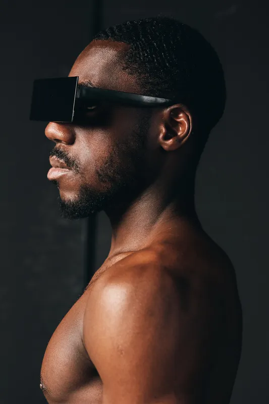 African man with dark skin and short hair wearing futuristic black sunglasses in creative conceptual visual style. Profile portrait with bare shoulders and modern fashion accessory.