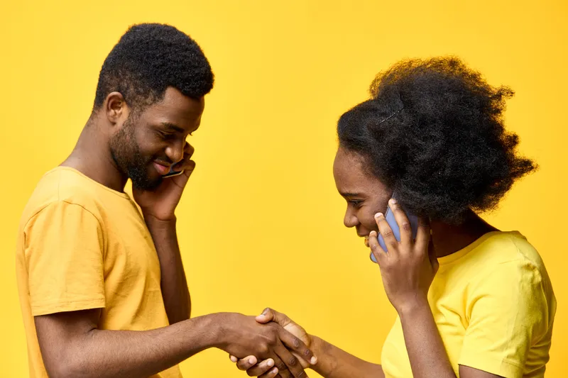 Two young African adults in casual yellow clothing engaging in a handshake while talking on mobile phones, set against a vibrant yellow background