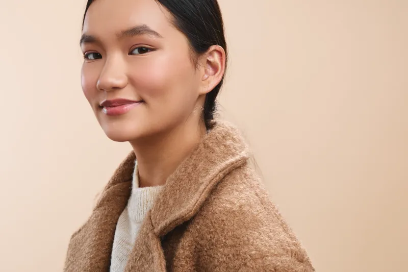 Asian woman with a warm smile stands in a soft beige studio, wearing a tan wool coat. She exudes calm confidence and approachable style, creating a friendly, contemporary mood.