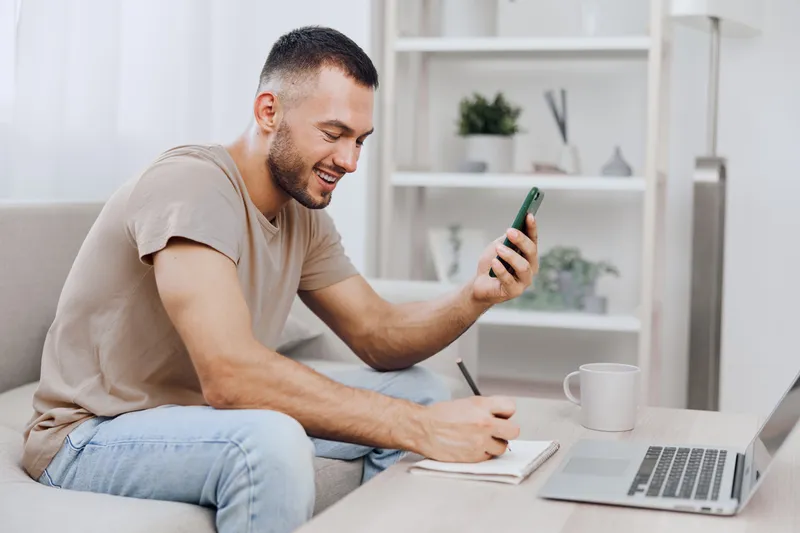 Smiling man engaged in work, writing notes while using smartphone, showcasing modern home office setup with bright and inviting atmosphere.