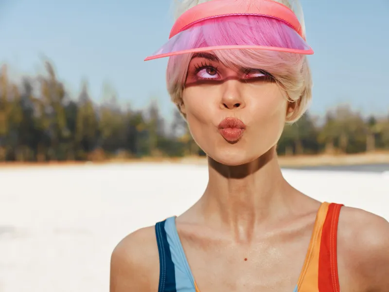 A young woman stands on a sunny beach, wearing a pink visor and colorful swimsuit. She blows a kiss toward the camera, radiating playful confidence and warm energy.