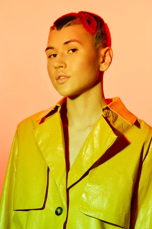 Young woman with red braided hair wearing a yellow leather jacket, posing confidently in warm orange lighting and minimalist background.
