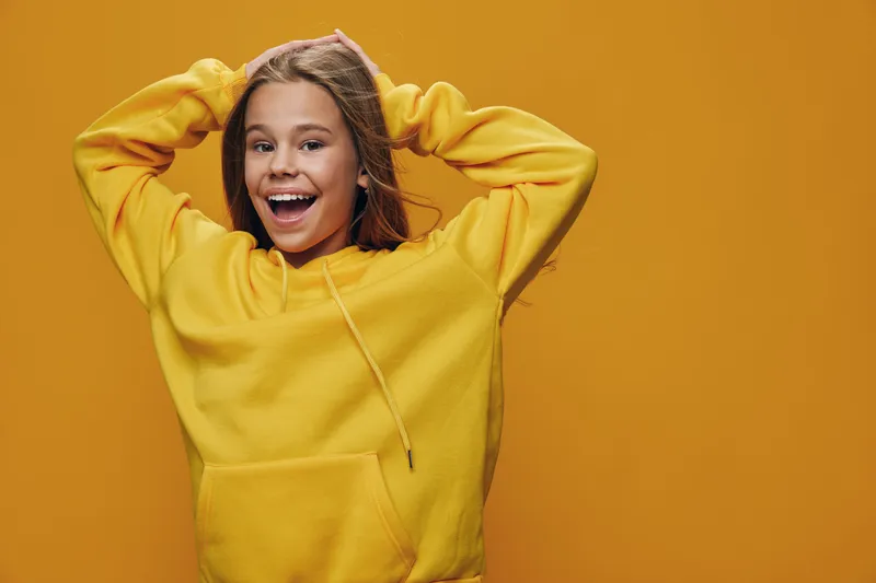 Girl with happy smile in yellow hoodie posing with hands behind head on orange background. Casual style, cheerful young person with confident expression indoors.