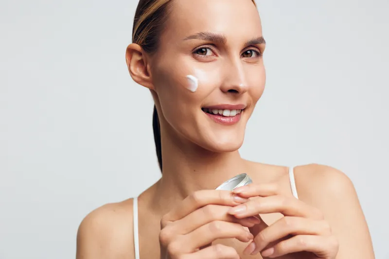 Smiling woman applies face cream on cheek holding open jar with natural skincare product isolated on white background with healthy skin and positive mood.