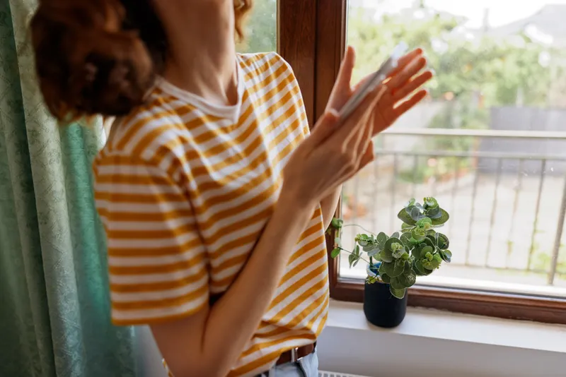 Casual woman indoors near a window, striped yellow shirt, clapping hands in a bright, relaxed home moment, soft natural light, green plant nearby, everyday lifestyle
