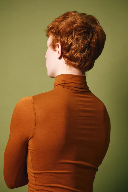 Person with red hair wearing brown turtleneck facing away standing against olive green background in studio portrait photography.