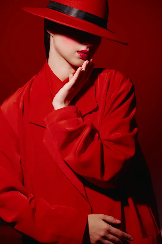 Fashionable woman wears vibrant red coat and hat, posing with creative hand gesture. High fashion beauty portrait with dramatic shadows and glossy red lipstick.