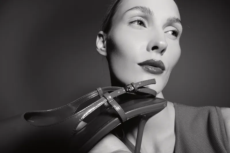 Caucasian woman with fair skin holds high heel shoe near face, looking sideways with soft studio lighting, fashion and style concept.