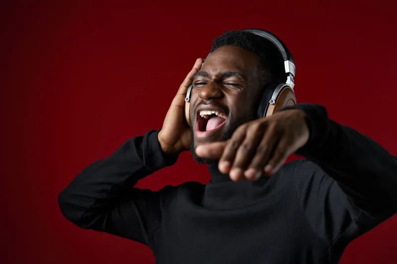 A joyful Black man in a black turtleneck and headphones singing passionately against a red background, showcasing the love for music and self expression
