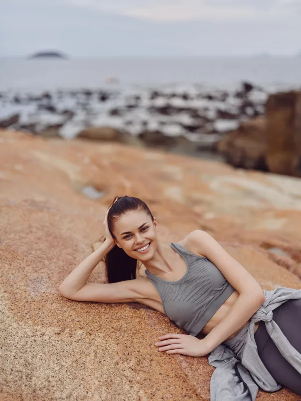 Young woman in gray athletic wear smiling while relaxing on rocky beach, embodying healthy lifestyle and summer vibes