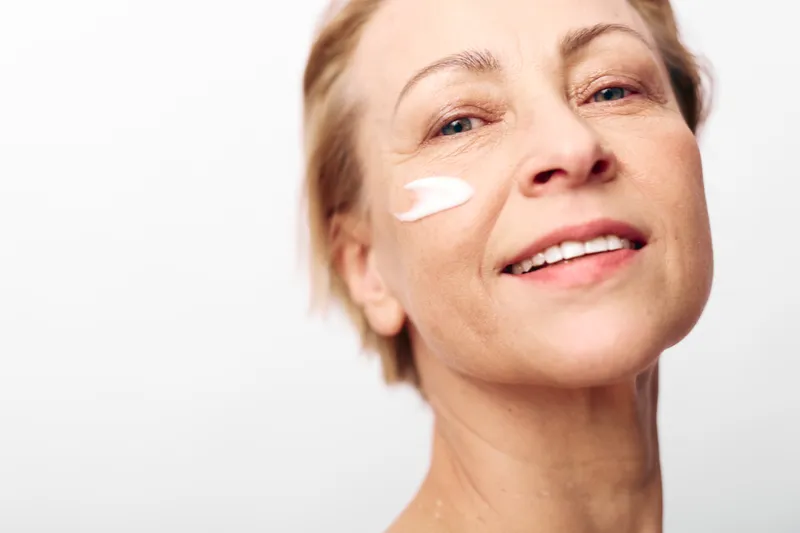 Mature woman with clear skin applies white face cream on cheek, smiling gently. Close-up portrait on plain background. Concept for skincare, beauty, anti aging, and self care.