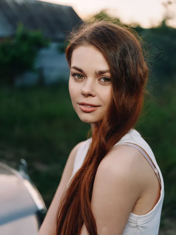 Young woman with long red hair sitting outdoors, wearing a casual white tank top, showcasing a serene expression in a natural green setting.