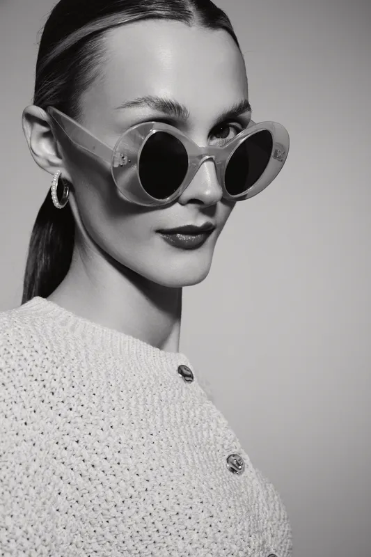 Stylish Caucasian woman with light skin wearing large round sunglasses and hoop earrings poses confidently in a textured sweater. Minimalist studio portrait in black and white.