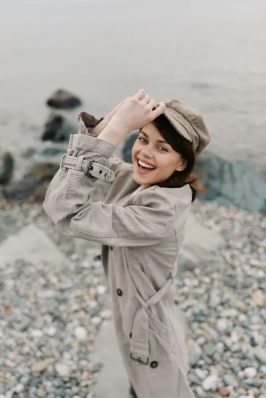 Smiling young woman in beige trench coat and matching hat poses on rocky beach, enjoying outdoor moment. Concept of casual fashion, happiness, and nature lifestyle by the sea.