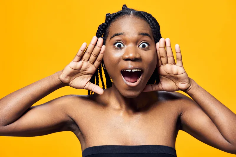 Excited young woman with braided hair expressing joy on a vibrant yellow background, celebrating positivity with an energetic pose and cheerful emotions