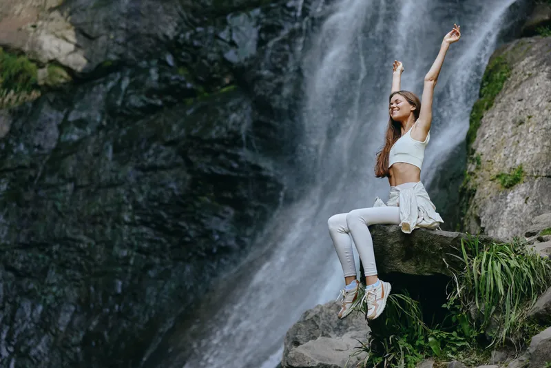 young woman enjoying waterfall, sitting on rock, wearing white activewear, peaceful nature scene, bright outdoor environment, relaxed mood