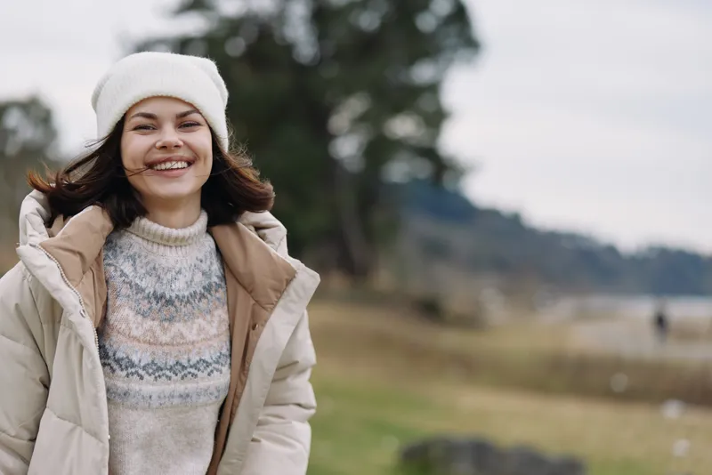 Woman in outdoor setting wearing a coat and knit sweater, smiling against a blurred landscape of grass and trees, winter attire and casual portrait in calm breeze and relaxed mood